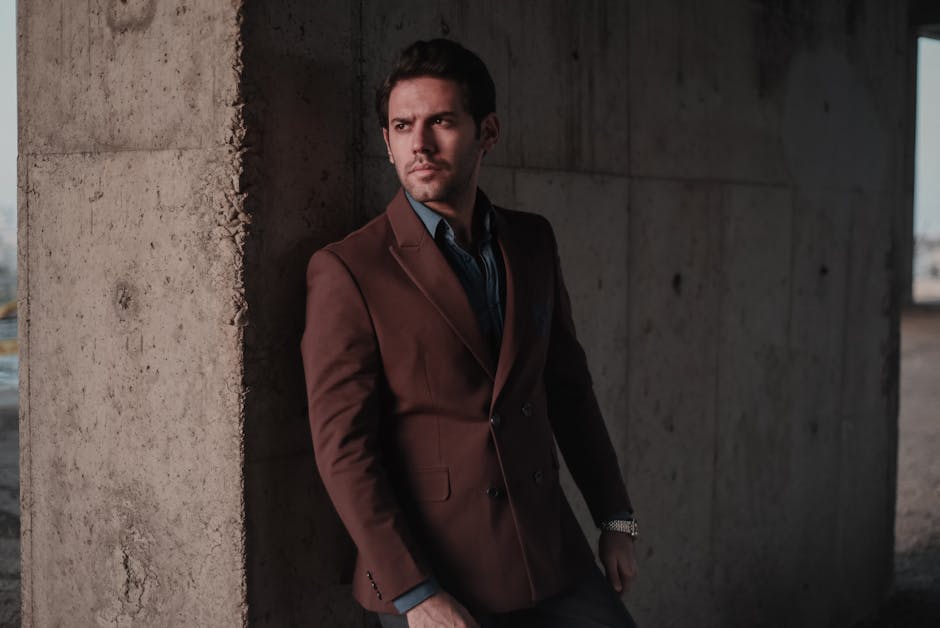 Elegant man in a brown suit posing outdoors against a concrete wall, showcasing fashion and style.