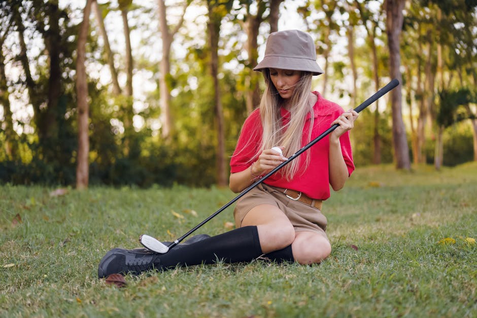 Young woman in fashionable attire with bucket hat sitting on grass, holding golf club.